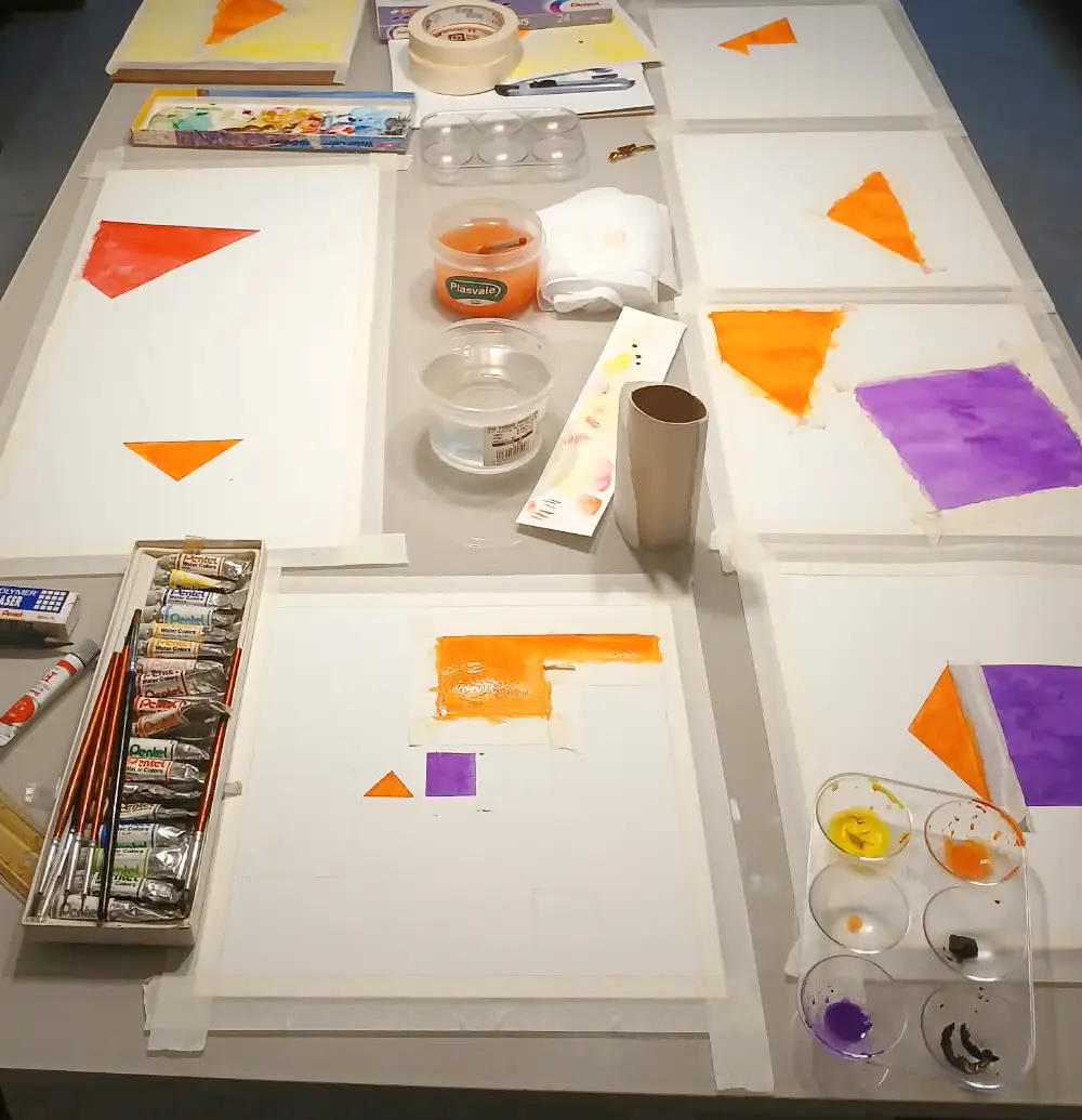 A table covered in taped watercolor sheets, brushes, pigments and glass mixing cups